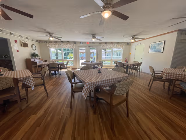 A dining room with multiple tables covered in checkered tablecloths, each surrounded by chairs. The room has wooden flooring, ceiling fans with lights, large windows letting in natural light, and a piano in the corner. The walls are light-colored with some decorations and a fire extinguisher near the door.