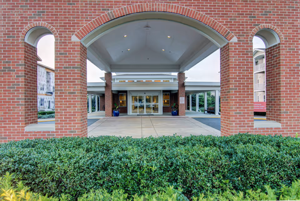 Entrance of a brick building with a covered driveway supported by brick columns, green shrubs in the foreground, and glass double doors leading inside.
