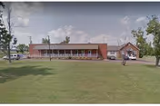 Single-story brick nursing home with a covered front porch, a few parked cars and a large grassy lawn under a partly cloudy sky.