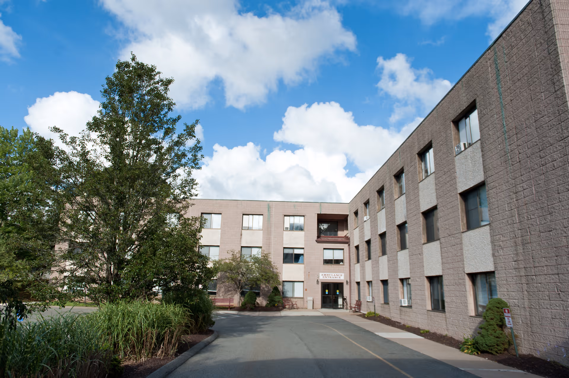Exterior view of a three-story healthcare facility building with multiple windows, a driveway leading to an ambulance entrance, and trees and shrubs surrounding the area under a partly cloudy blue sky.