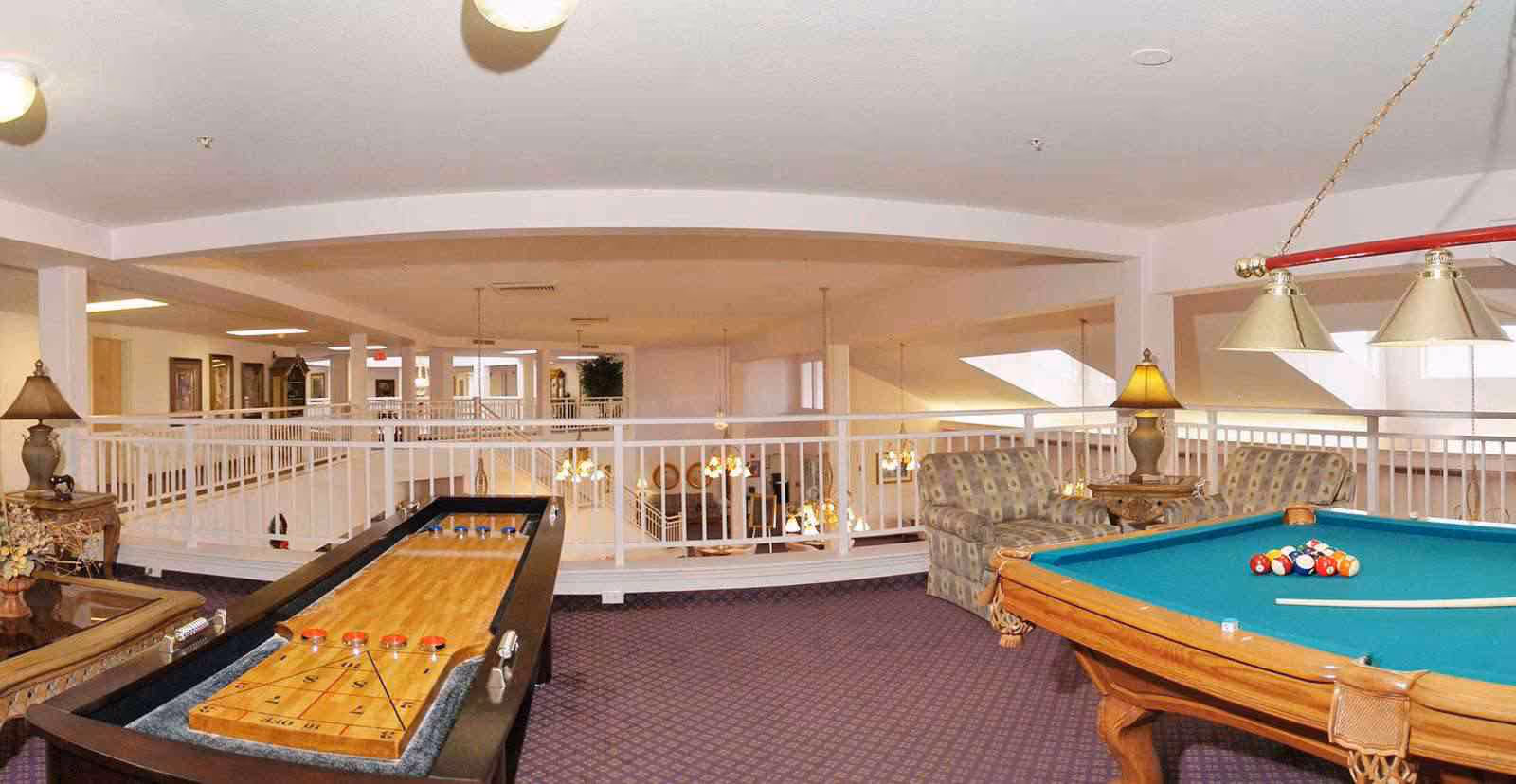 Upstairs common area with a pool table, shuffleboard, upholstered chairs and lamps overlooking a lower-level railing.