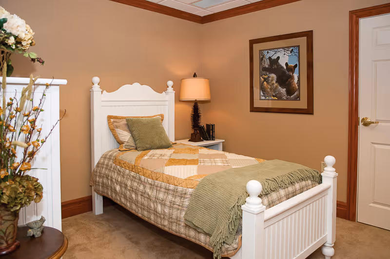 A cozy bedroom with a single white wooden bed featuring a patchwork quilt and green throw blanket. There are two pillows on the bed, one beige and one green. A nightstand beside the bed holds a lamp with a beige shade and some books. A framed picture of bears hangs on the beige wall above the nightstand. The room has beige walls with wooden trim and a closed white door with a brass handle.