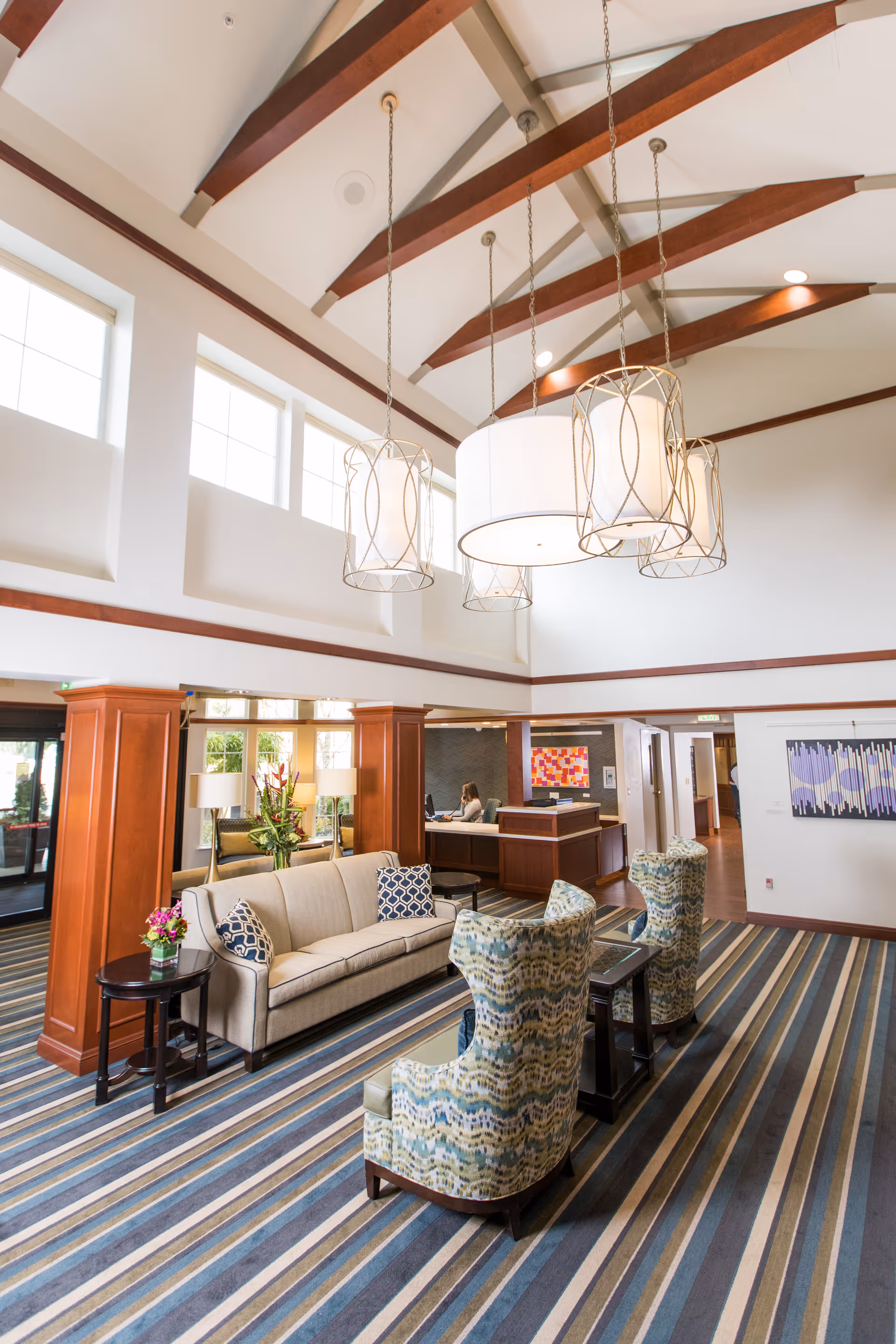 Bright high-ceilinged lobby with hanging chandeliers, a sofa and patterned armchairs on a striped carpet in front of a reception desk.