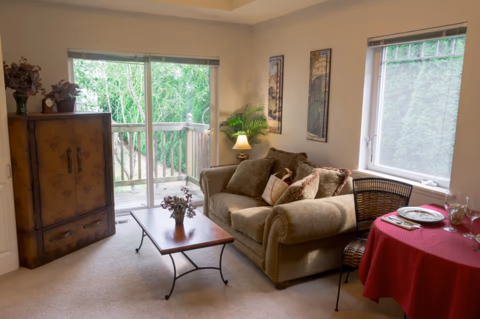 Cozy living room area with a beige sofa adorned with multiple cushions, a wooden coffee table with a small plant, a wooden cabinet with decorative items on top, a small round dining table covered with a red tablecloth set with plates and glasses, and large windows letting in natural light with a view of green trees outside.