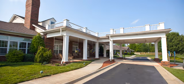 Covered porte-cochere entrance with white columns at the front of a brick senior living building and a paved driveway under a blue sky.