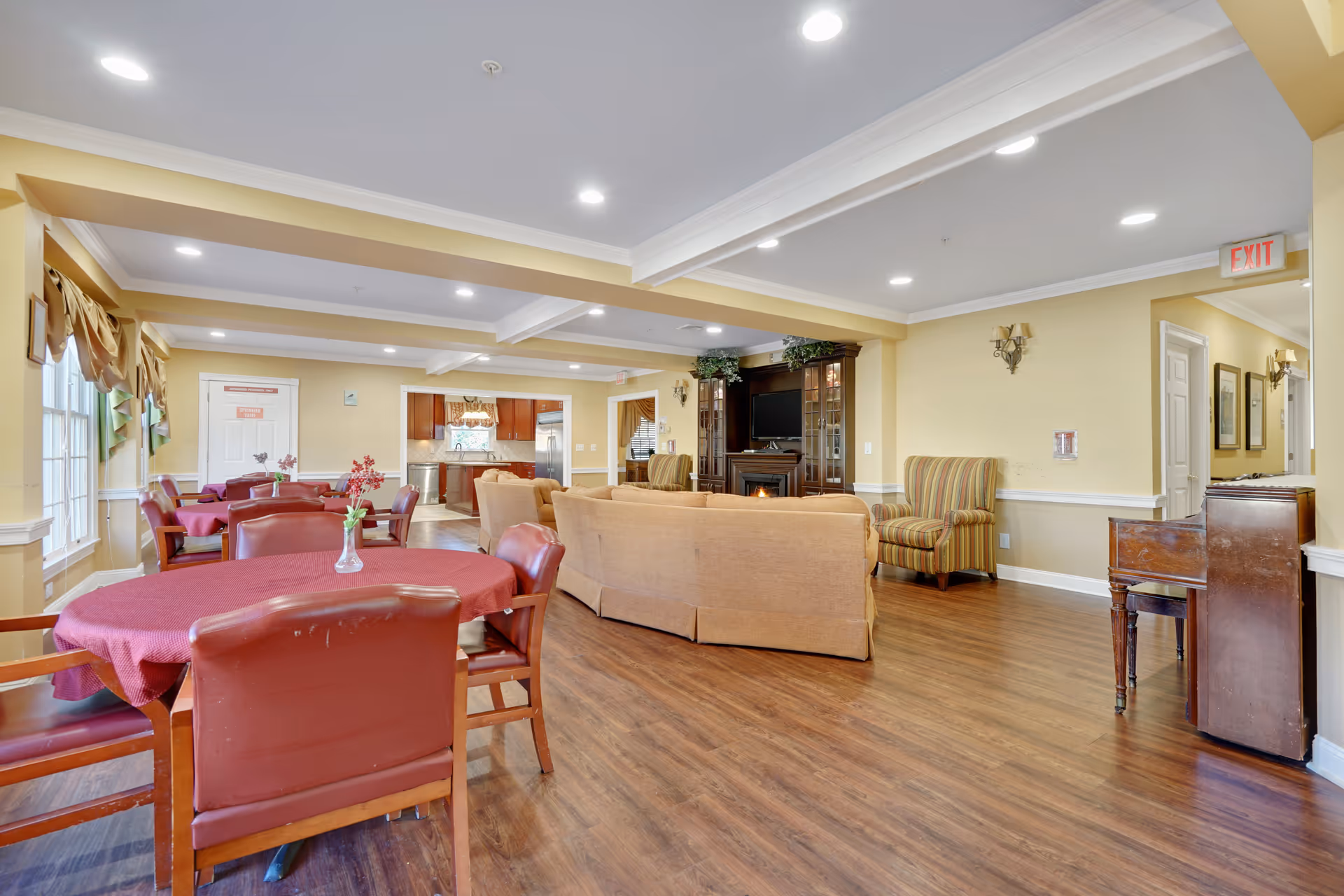 A spacious, well-lit common area in a senior living facility featuring round tables with red tablecloths and chairs, a seating area with beige sofas and a striped armchair, a fireplace with a TV above it, and a view into a kitchen area in the background. The room has wooden flooring, yellow walls, and recessed ceiling lights.