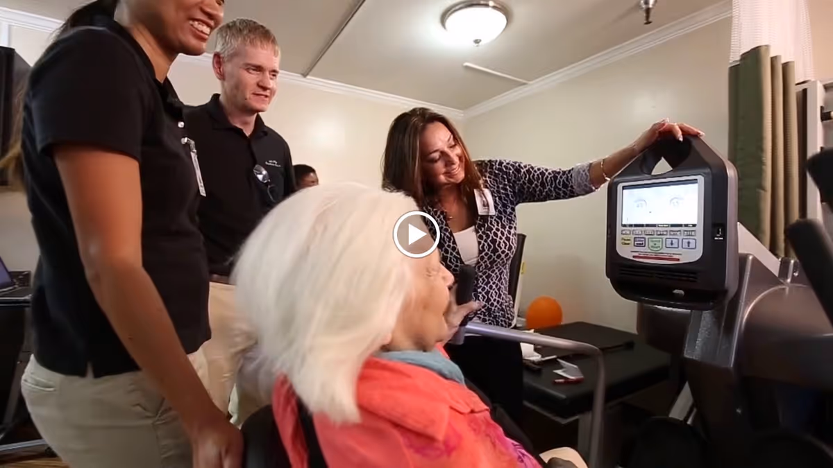 An elderly woman with white hair in a wheelchair is interacting with a touchscreen device, while three staff members stand nearby watching and smiling in a well-lit room.