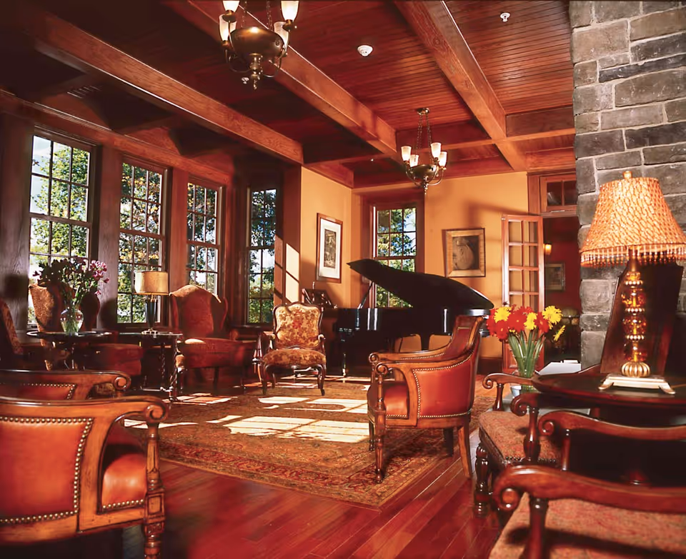 Warm, wood-paneled living room with a grand piano, leather armchairs, large windows, exposed ceiling beams, and a stone fireplace.
