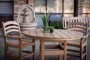 Outdoor patio area with a round wooden table and two wooden chairs. On the table are three small potted plants. In the background, there is a decorative stone water fountain mounted on a brick wall and a window with blinds.