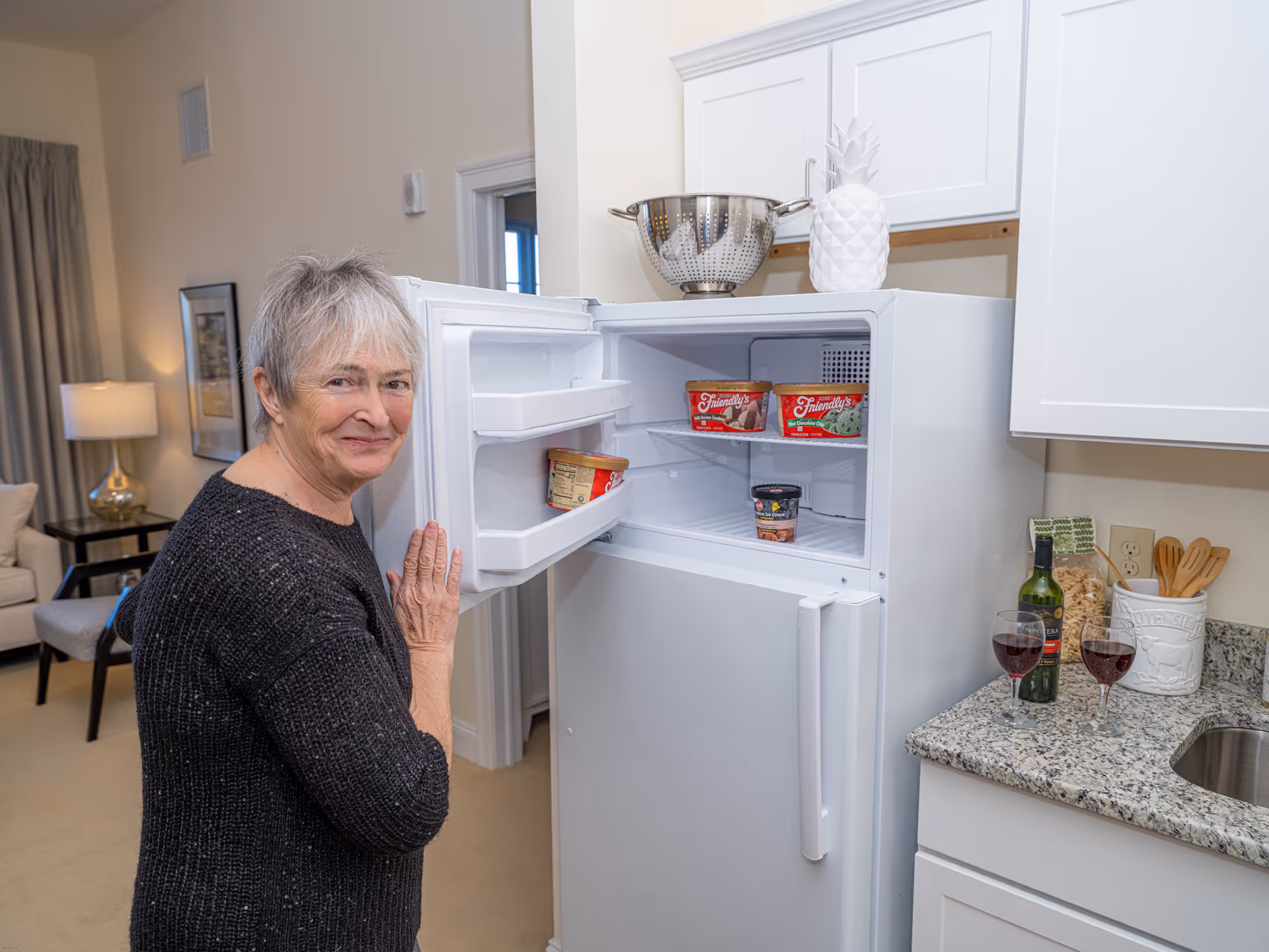 An elderly woman with short gray hair and a black sweater stands next to an open white refrigerator freezer in a kitchen. The freezer contains several ice cream containers. The kitchen has white cabinets, a granite countertop with two glasses of red wine, a bottle of wine, and kitchen utensils. In the background, a living room area with a lamp, chair, and framed picture is visible.