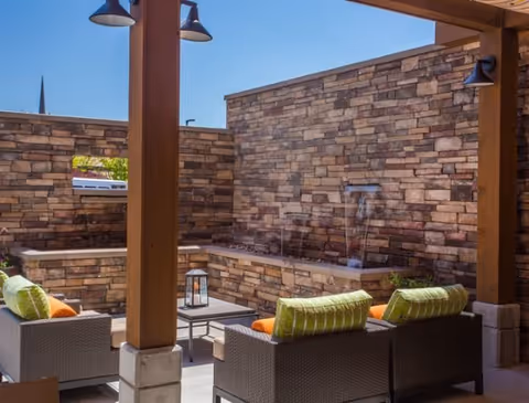Outdoor patio area with wicker furniture including chairs with green and orange cushions, a small table with a lantern, and a stone wall with water features under a clear blue sky.