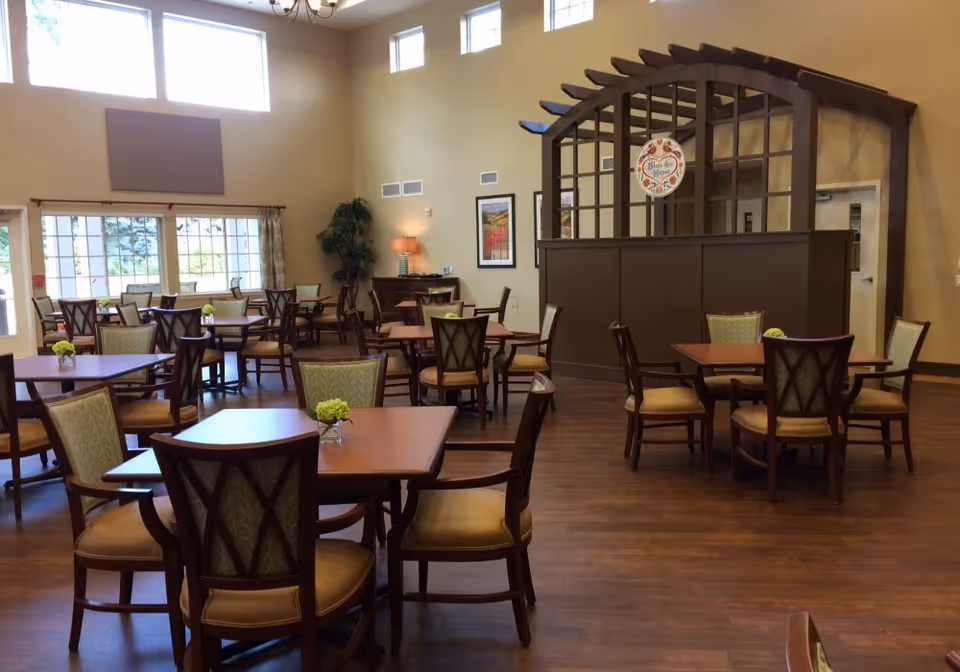 Bright dining room with multiple wooden tables and upholstered chairs, a decorative wooden pergola divider, and high windows.