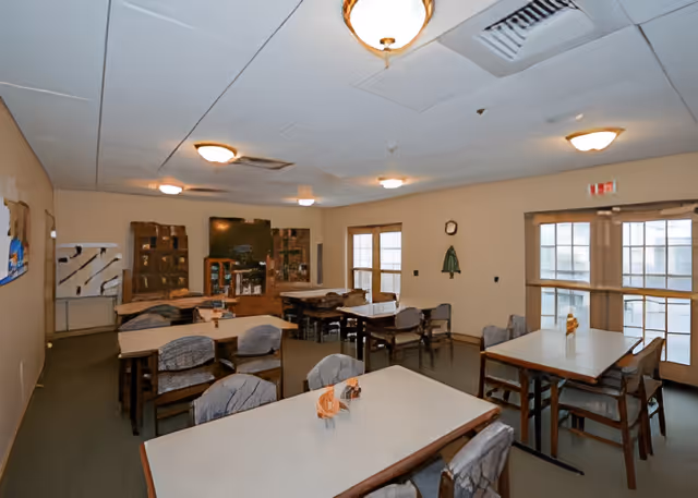 A dining room with several tables and chairs arranged neatly. The room has beige walls, multiple ceiling lights, and large windows letting in natural light. There is a television mounted on the far wall with shelves and cabinets below it. The tables have small decorative items placed on them.
