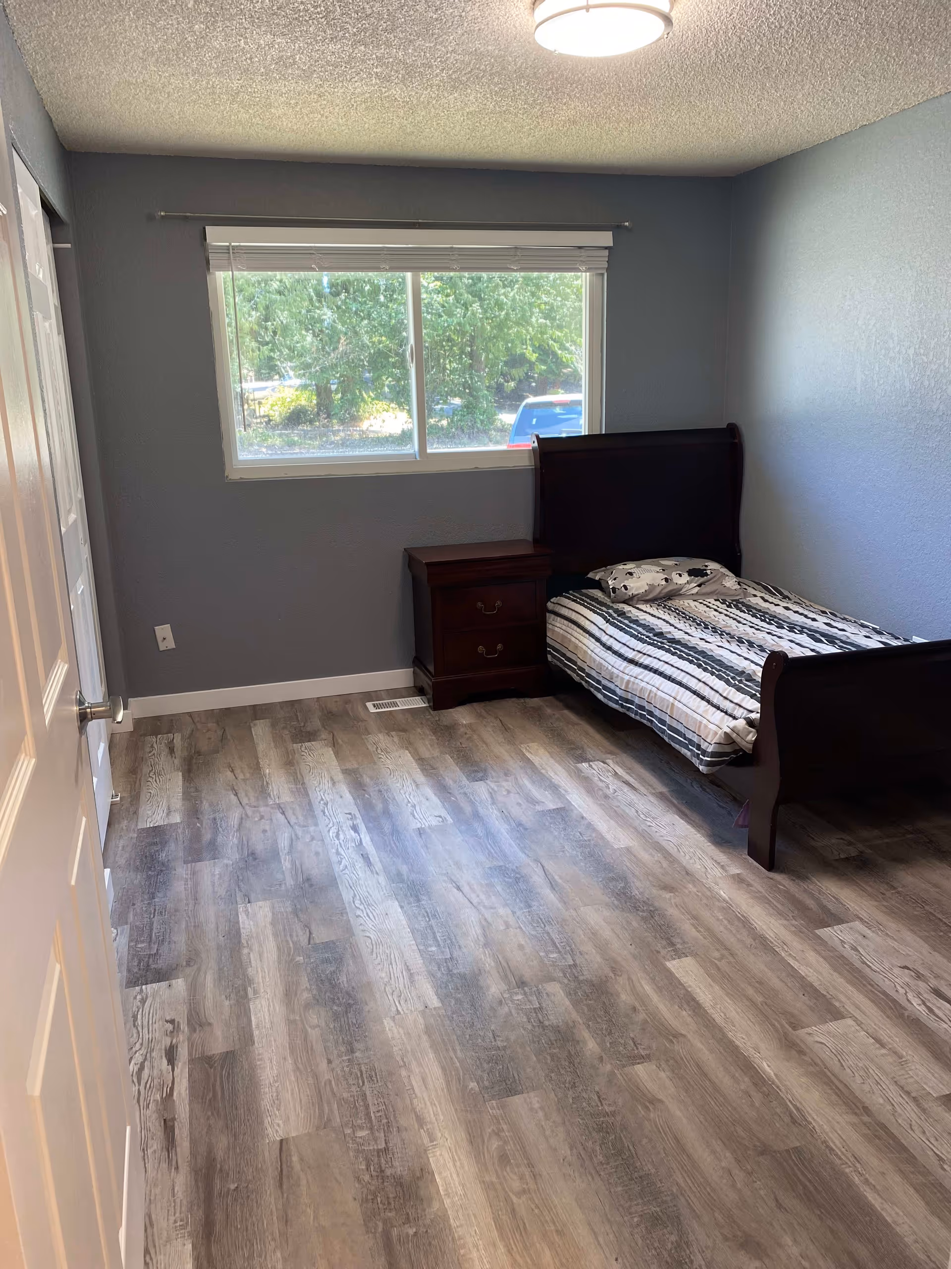Small bedroom with a single bed, nightstand, window, and gray wood-look flooring.