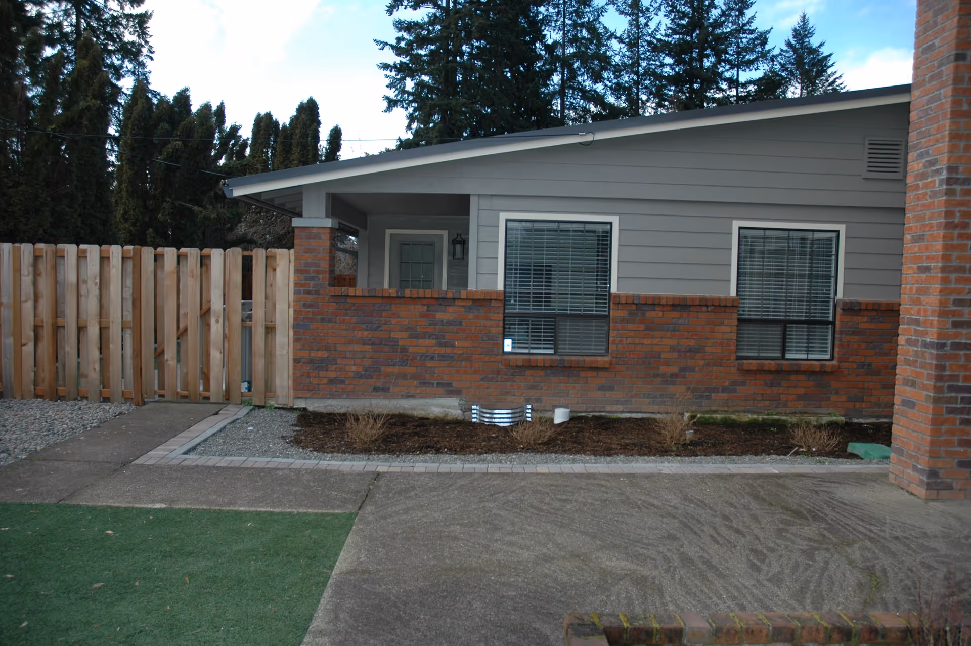 Exterior view of a single-story building with gray siding and a brick lower half. There are two windows with white blinds and a door under a covered porch area. A wooden fence is on the left side, and there is a concrete pathway and some landscaping with mulch and small bushes in front of the building. Tall evergreen trees are visible in the background under a partly cloudy sky.