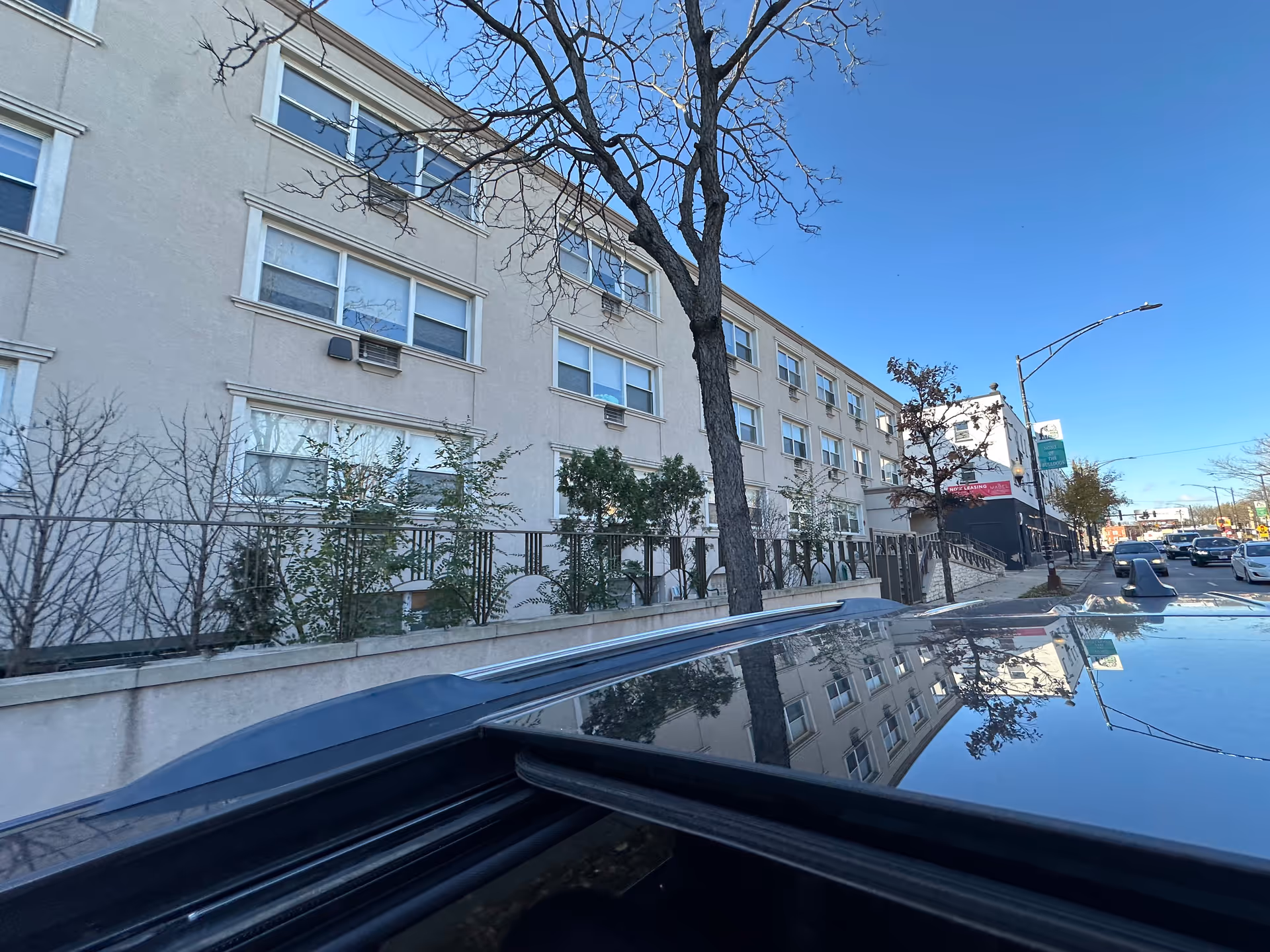 Exterior view of a multi-story beige building with multiple windows, some with air conditioning units. There are leafless trees and shrubs along a fenced area in front of the building. The sky is clear and blue, and part of a car roof is visible in the foreground reflecting the building and trees.