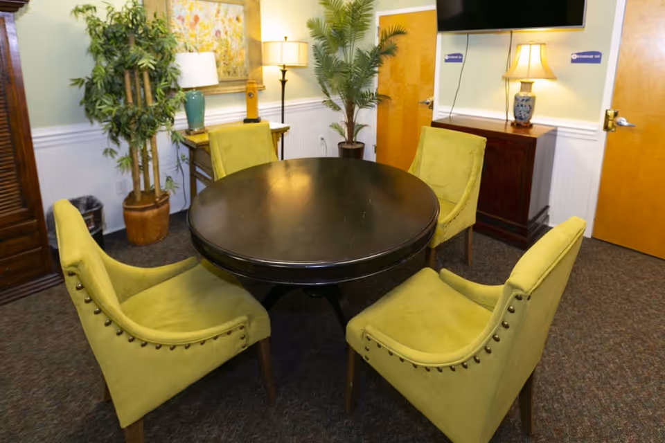 A small interior room with a round black table surrounded by four green upholstered chairs. The room has two wooden doors, a wooden cabinet with a lamp and a TV mounted above it, a side table with a lamp and a decorative vase, two potted plants, and a framed floral painting on the wall.