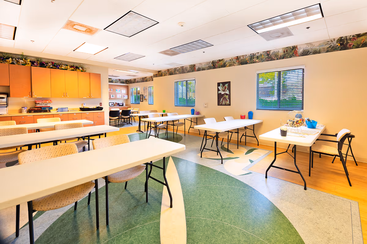 A bright, spacious room with multiple white folding tables and beige cushioned chairs arranged in rows. The room has a green and beige patterned floor, light yellow walls with floral wallpaper border near the ceiling, and two windows with blinds letting in natural light. There are cabinets along one wall with various items on the countertop, and a small table with bingo equipment on the right side.