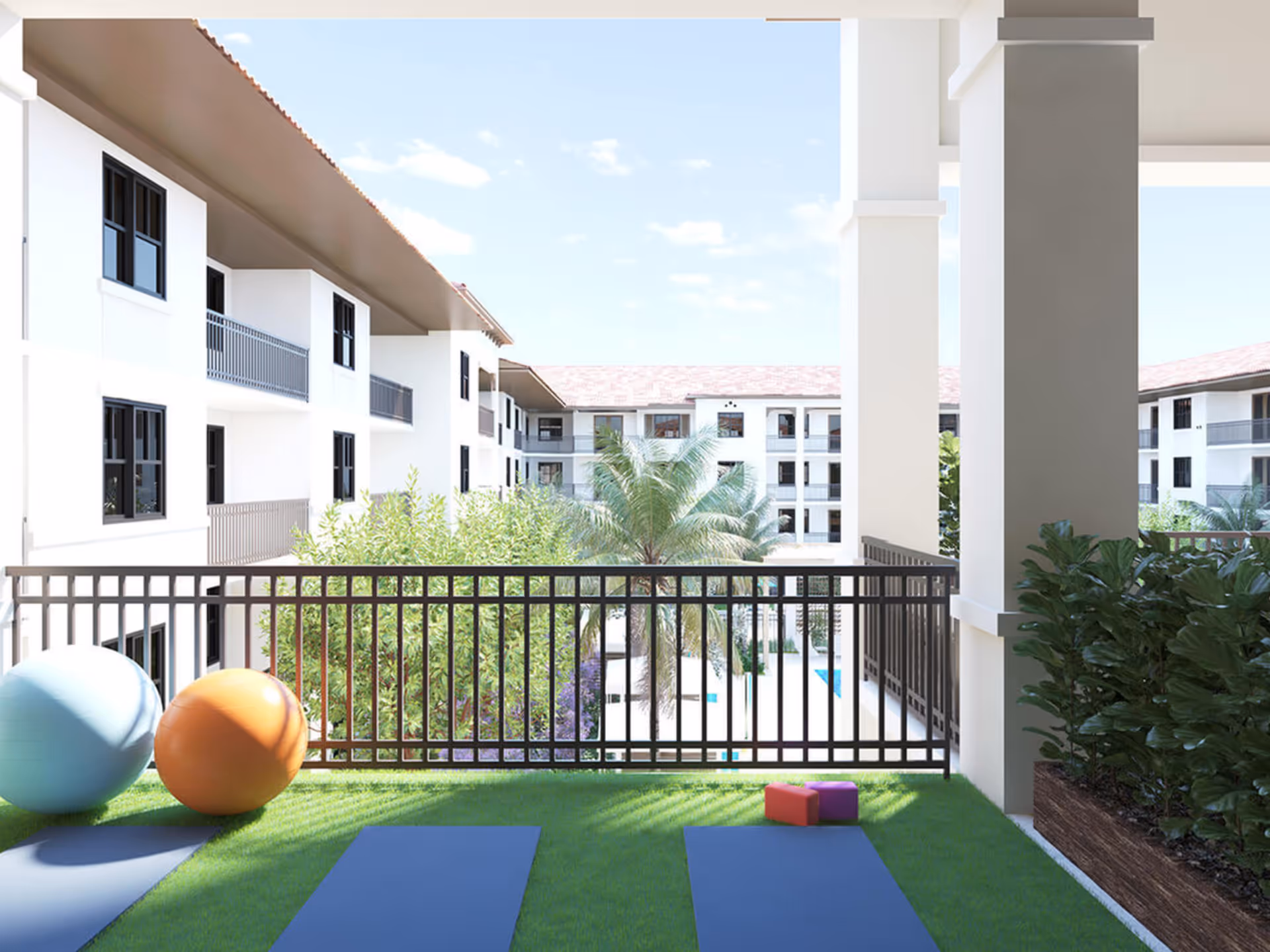 Outdoor balcony area with two exercise mats, two large exercise balls, and two small yoga blocks. The balcony overlooks a courtyard with palm trees and a swimming pool, surrounded by a multi-story white building with balconies and windows under a clear blue sky.