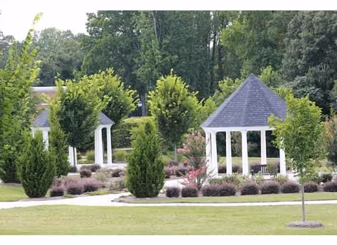 A landscaped outdoor area featuring two white gazebos with gray roofs surrounded by green trees, shrubs, and neatly trimmed grass. There are walking paths winding through the garden area.