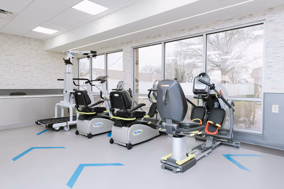 A rehabilitation exercise room with several pieces of fitness equipment including two NuStep recumbent cross trainers and a rowing machine, positioned near large windows that let in natural light. The room has light-colored walls and flooring with blue directional markings on the floor.