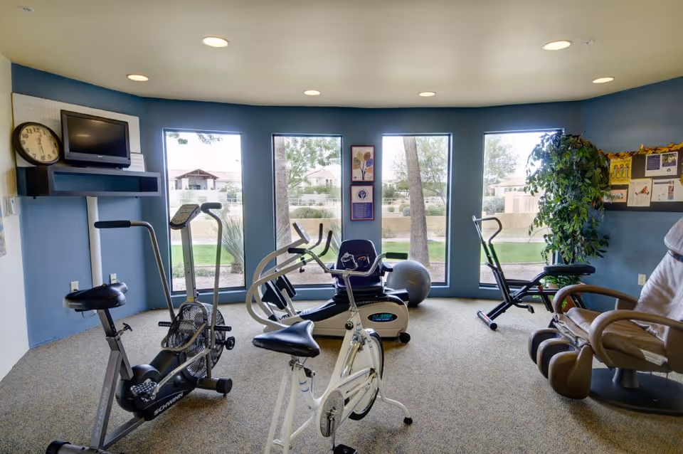 A small exercise room with several stationary bikes and exercise equipment, large windows showing an outdoor view with trees and houses, a clock and a TV mounted on the wall, and a comfortable chair near a bulletin board with notices.