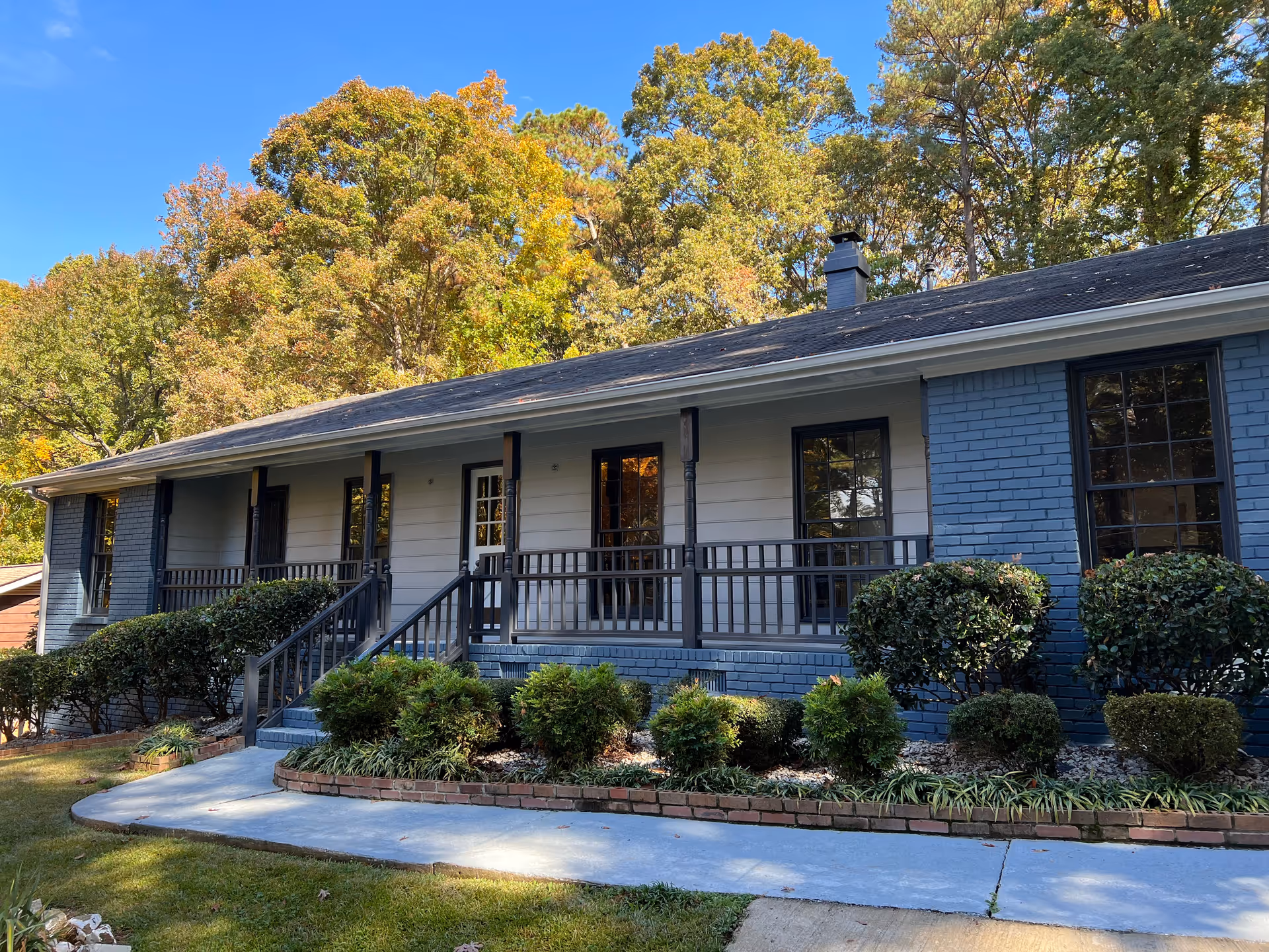 Exterior view of a single-story residential building with a front porch, black railings, and steps leading up to the entrance. The building has a combination of blue brick and white siding, surrounded by neatly trimmed bushes and trees with autumn foliage under a clear blue sky.