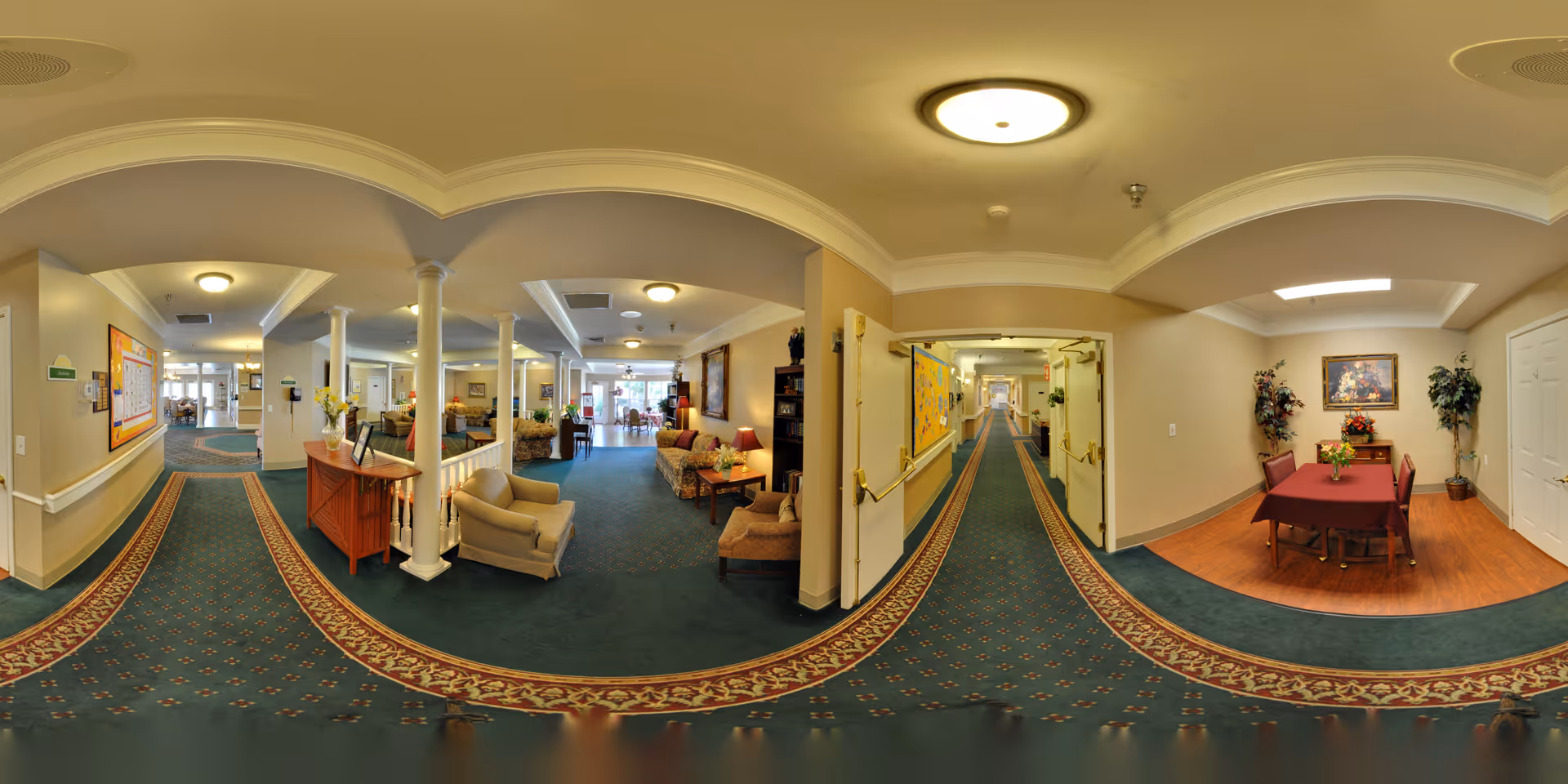 A spacious and well-lit senior living facility interior with a long carpeted hallway featuring a patterned border. To the left, there is a seating area with armchairs and a small table with flowers. To the right, there is a small dining area with a table covered by a red tablecloth and four chairs, decorated with plants and a framed painting on the wall. The ceiling has recessed lighting and decorative molding.