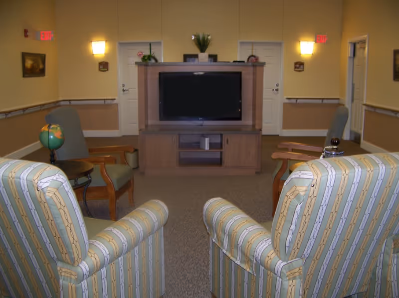 Communal living room with patterned armchairs facing a television on a wooden media cabinet.