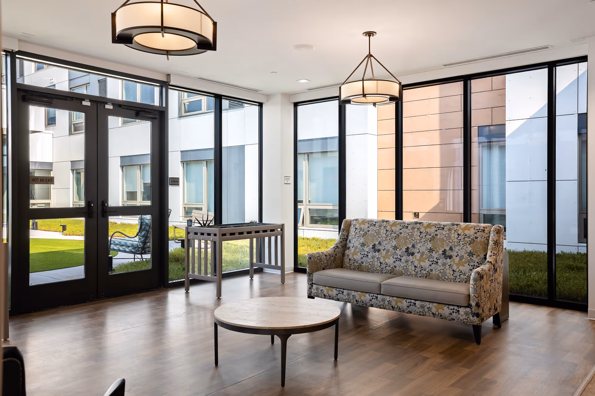 Sunlit lounge with floor-to-ceiling windows, a patterned sofa, round coffee table, and glass doors opening to a courtyard.