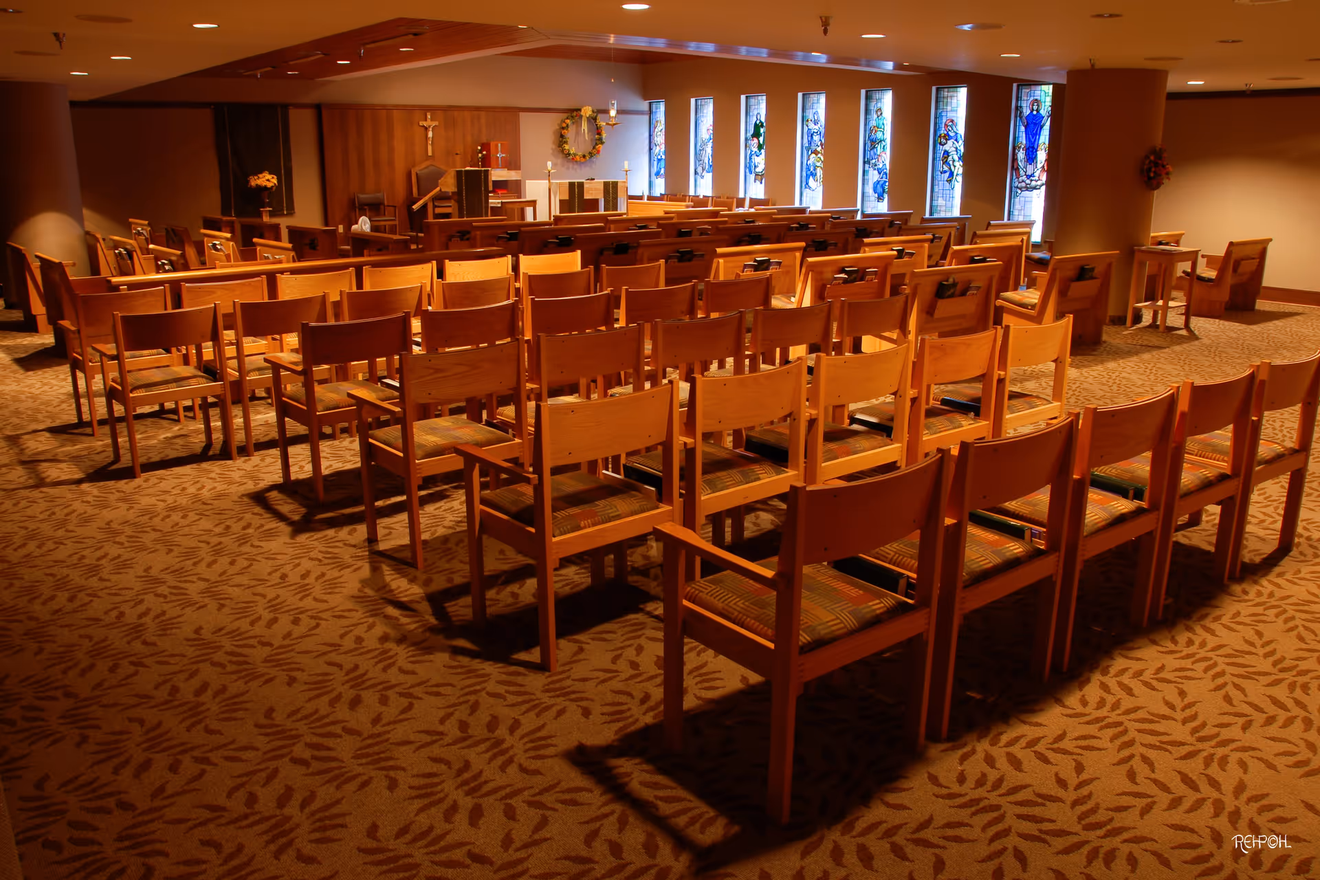 Empty chapel-style room with rows of wooden chairs facing an altar and stained-glass windows.