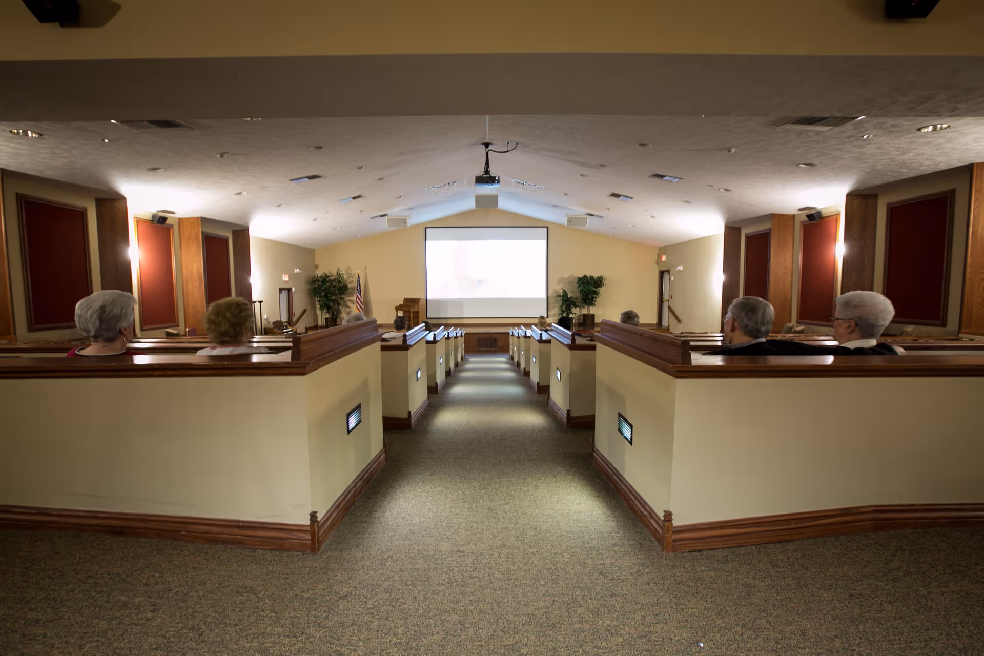 Interior view of a small theater or presentation room with several elderly people seated in wooden pew-style benches facing a large projection screen at the front. The room has beige walls, red acoustic panels, and soft lighting along the walls.