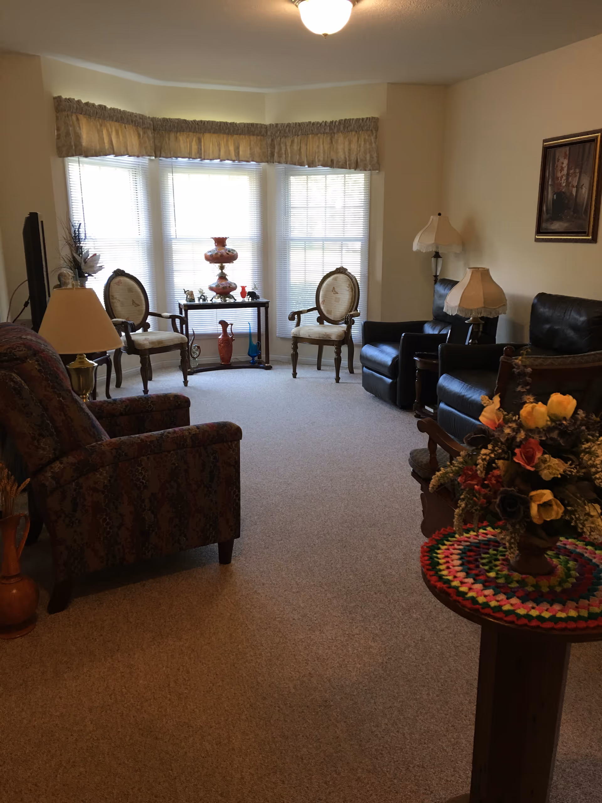 A cozy living room with beige carpet and cream-colored walls. The room features a patterned armchair, two vintage-style upholstered chairs, and two black leather armchairs. There are two table lamps with white shades, a small table with decorative vases and figurines in front of a large bay window with blinds and a valance. A round table with a colorful crocheted doily and a floral arrangement is in the foreground. A framed painting hangs on the wall.