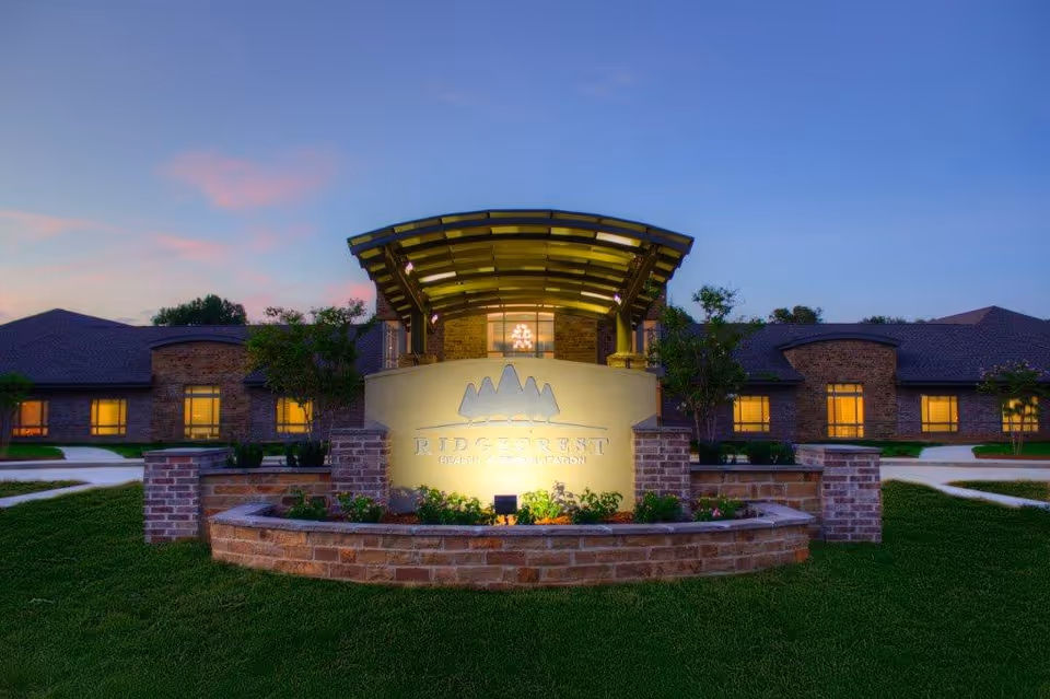 Exterior view of Ridgecrest Health And Rehabilitation facility at dusk, showing a well-lit entrance sign with landscaping and a building with multiple windows in the background.
