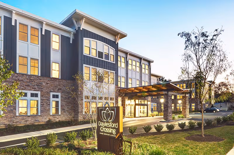 Exterior view of Daylesford Crossing senior living facility at dusk, showing a modern three-story building with large windows, stone and siding facade, a covered entrance, landscaped bushes and trees, and a sign with the facility name.