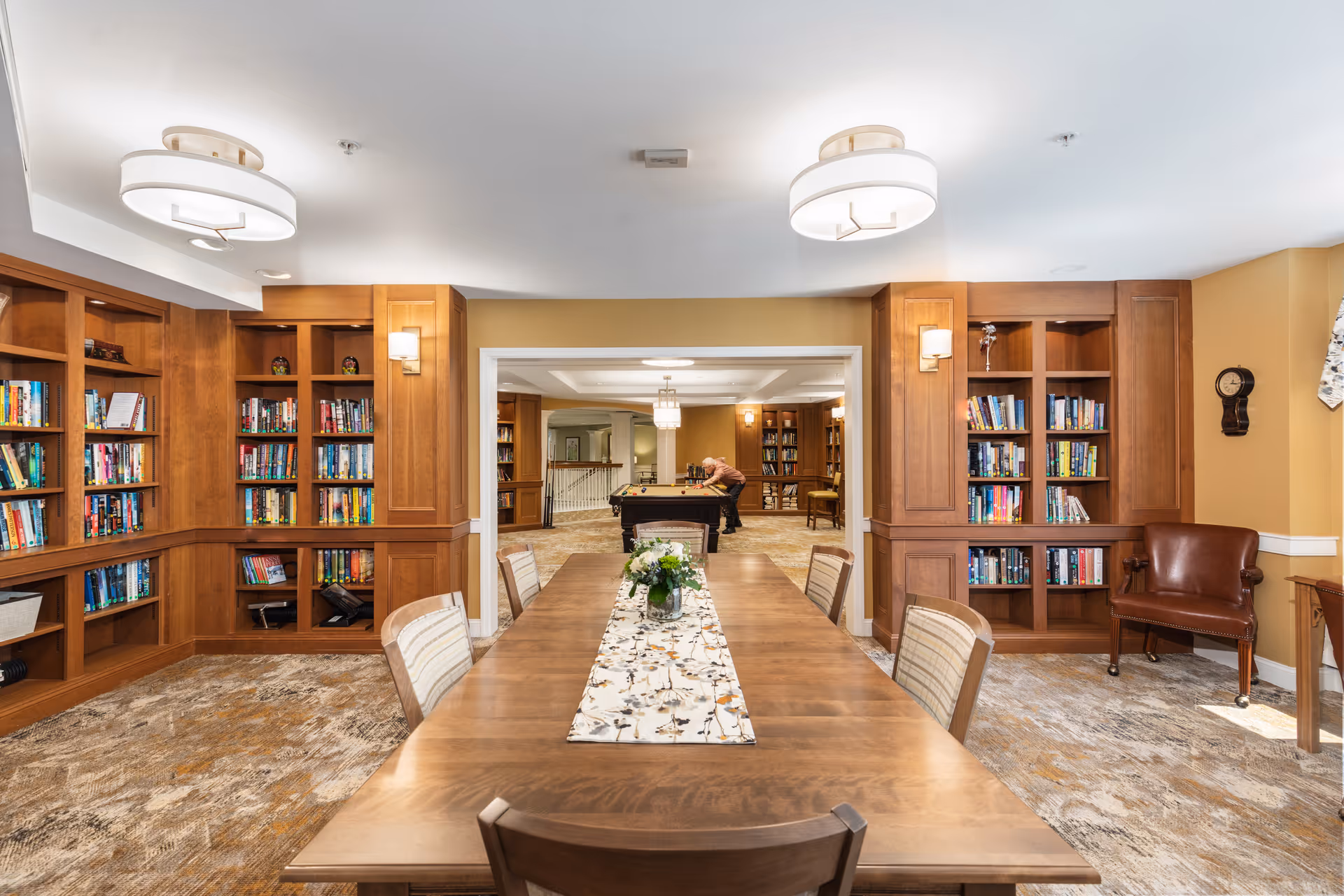 Communal library and game room with a long wooden table and chairs, built-in bookshelves, and a pool table visible in the adjoining room.