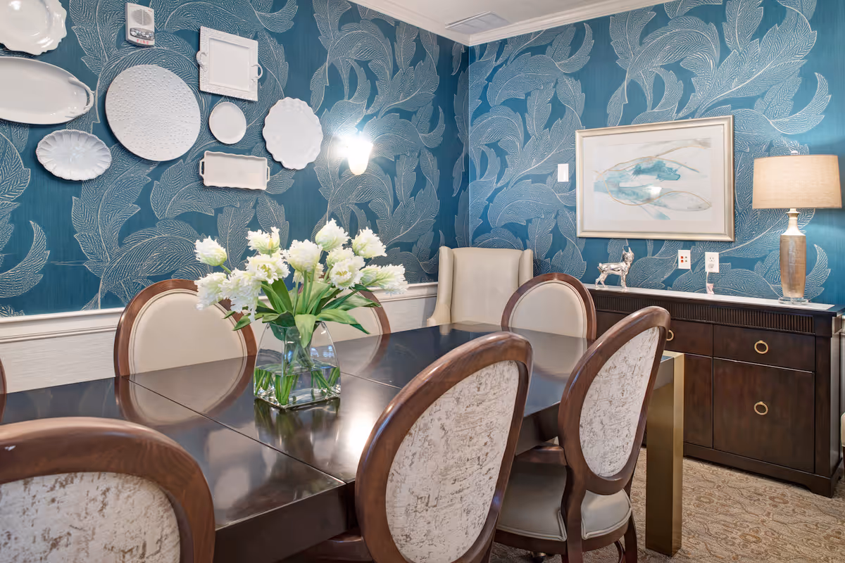 A dining room with a dark wooden table surrounded by upholstered chairs. A glass vase with white flowers is placed on the table. The walls are decorated with blue wallpaper featuring a large leaf pattern and adorned with white decorative plates. A sideboard with a lamp, a small silver dog figurine, and a framed abstract artwork is against the wall.