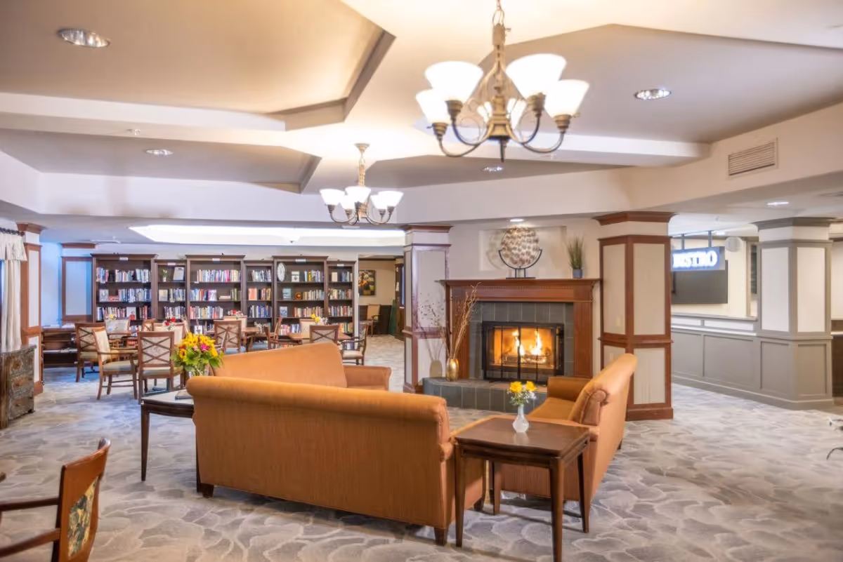 A cozy senior living facility common area with a lit fireplace, two orange sofas facing each other, a wooden side table with a small vase of flowers, bookshelves filled with books, multiple tables and chairs, and warm ceiling lights.