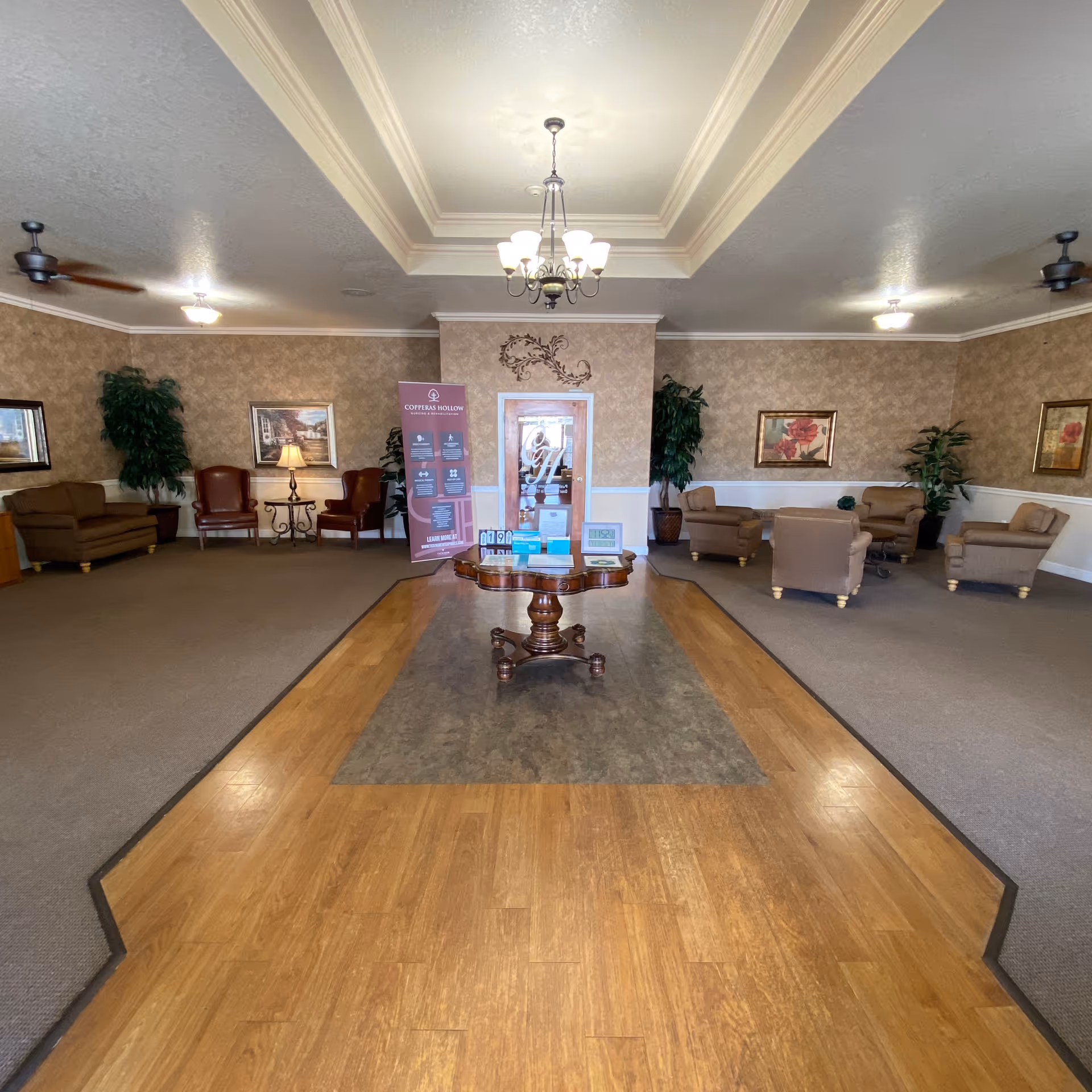 A spacious and well-lit common area in a senior living facility featuring a central wooden table with informational materials, surrounded by comfortable armchairs and sofas. The room has beige walls with framed artwork, potted plants, ceiling fans, and a chandelier hanging from a tray ceiling. A door with the facility's logo and a standing banner are visible at the back.