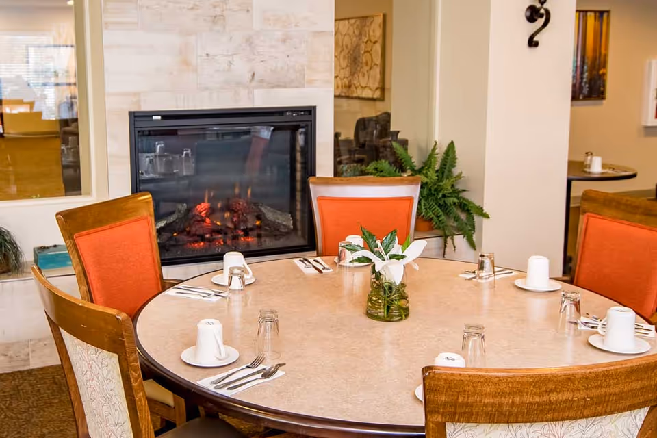 A round dining table set for six with white cups, glasses, and silverware, positioned in front of a modern electric fireplace with a warm fire. The chairs around the table have wooden frames with orange and patterned upholstery. A small vase with green leaves and white flowers is placed in the center of the table. The room has light-colored walls and carpeted flooring.