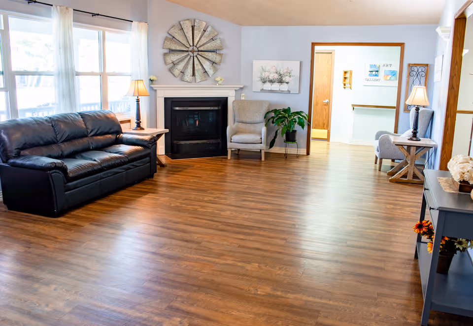 A bright and spacious living room with wooden flooring, a black leather sofa, two beige armchairs, a fireplace with a decorative metal windmill above it, a potted plant, table lamps on side tables, and wall art including a floral painting and art gallery signs in the background.