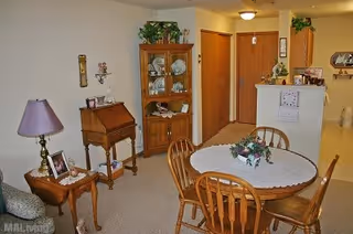 A cozy dining area in an apartment featuring a round wooden table with a white lace tablecloth and a floral centerpiece. Four wooden chairs surround the table. To the left, there is a small side table with a purple lamp and framed photos. Behind the dining table, a wooden cabinet displays decorative items and dishes. The background shows a hallway with wooden doors and a partial view of a kitchen counter with plants and more decor.