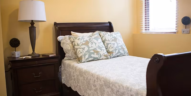 A neatly made single bed with a wooden headboard and footboard, adorned with patterned pillows and a light-colored bedspread. Next to the bed is a wooden nightstand with a lamp, a small decorative plant, and a book. A window with blinds allows natural light into the room.