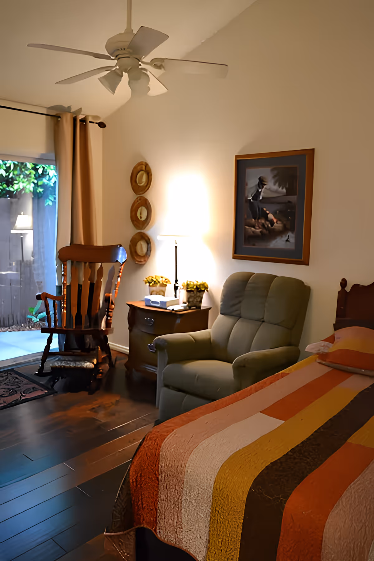 Cozy bedroom with a striped quilted bed, green recliner, wooden rocking chair, nightstand with a lamp, and a ceiling fan.