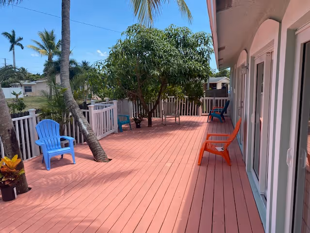 Outdoor wooden deck area with several colorful chairs including blue and orange ones, surrounded by a white fence and palm trees under a clear blue sky.