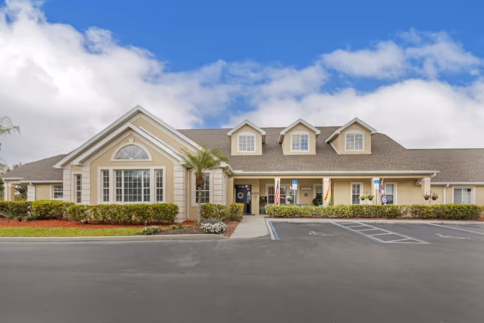 Front exterior view of a single-story beige building with a gabled roof, three dormer windows, and a large window with an arched top. There are small bushes and landscaping in front, a paved parking area with handicap parking spaces, and flags near the entrance under a covered porch.