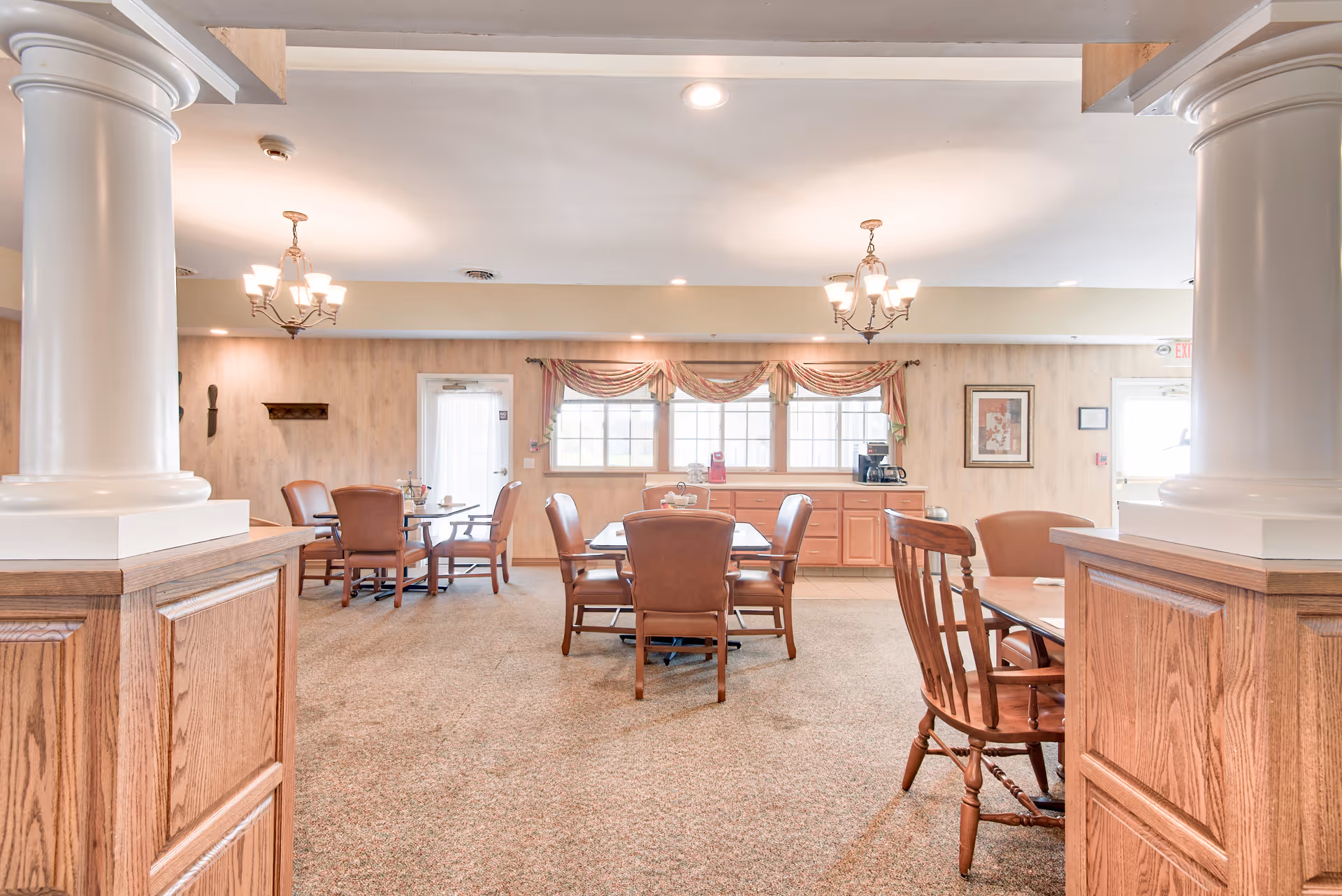 A well-lit dining room with multiple wooden tables and chairs arranged on a carpeted floor. The room features two large white columns in the foreground, chandeliers hanging from the ceiling, and windows with decorative valances letting in natural light. There is a sideboard with a coffee maker and other items against the far wall, along with framed artwork and a door leading outside.