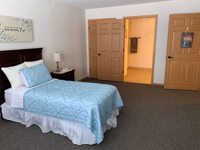 Tidy single bedroom with a twin bed covered in blue bedding, a nightstand with a lamp, and doors leading to a bathroom and closet.