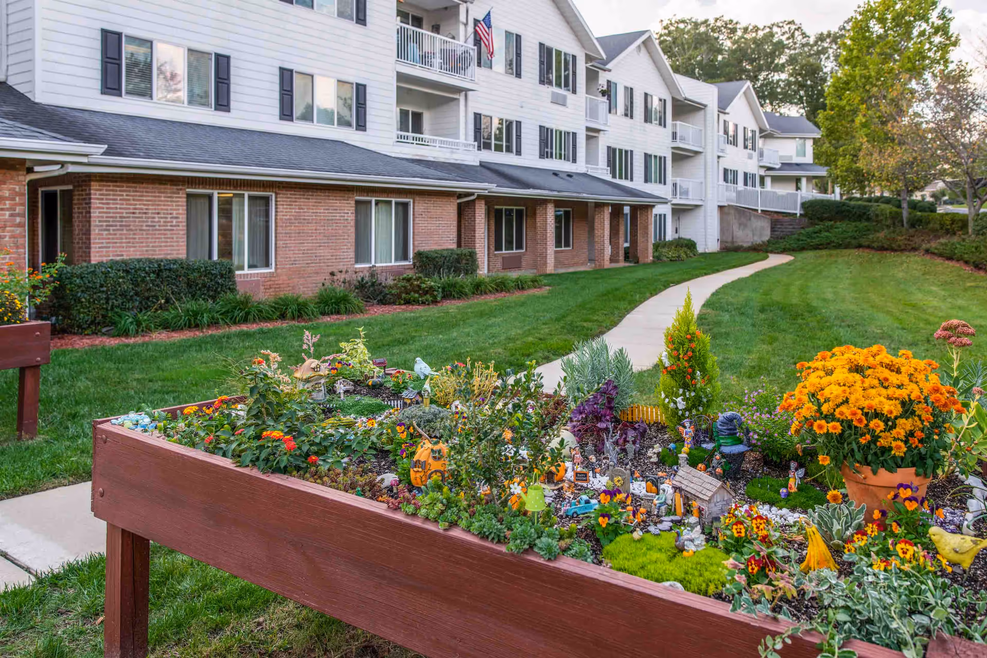 Raised garden bed filled with various colorful flowers and small decorative items in front of a multi-story residential building with brick and white siding exterior, surrounded by green grass and a paved walkway.