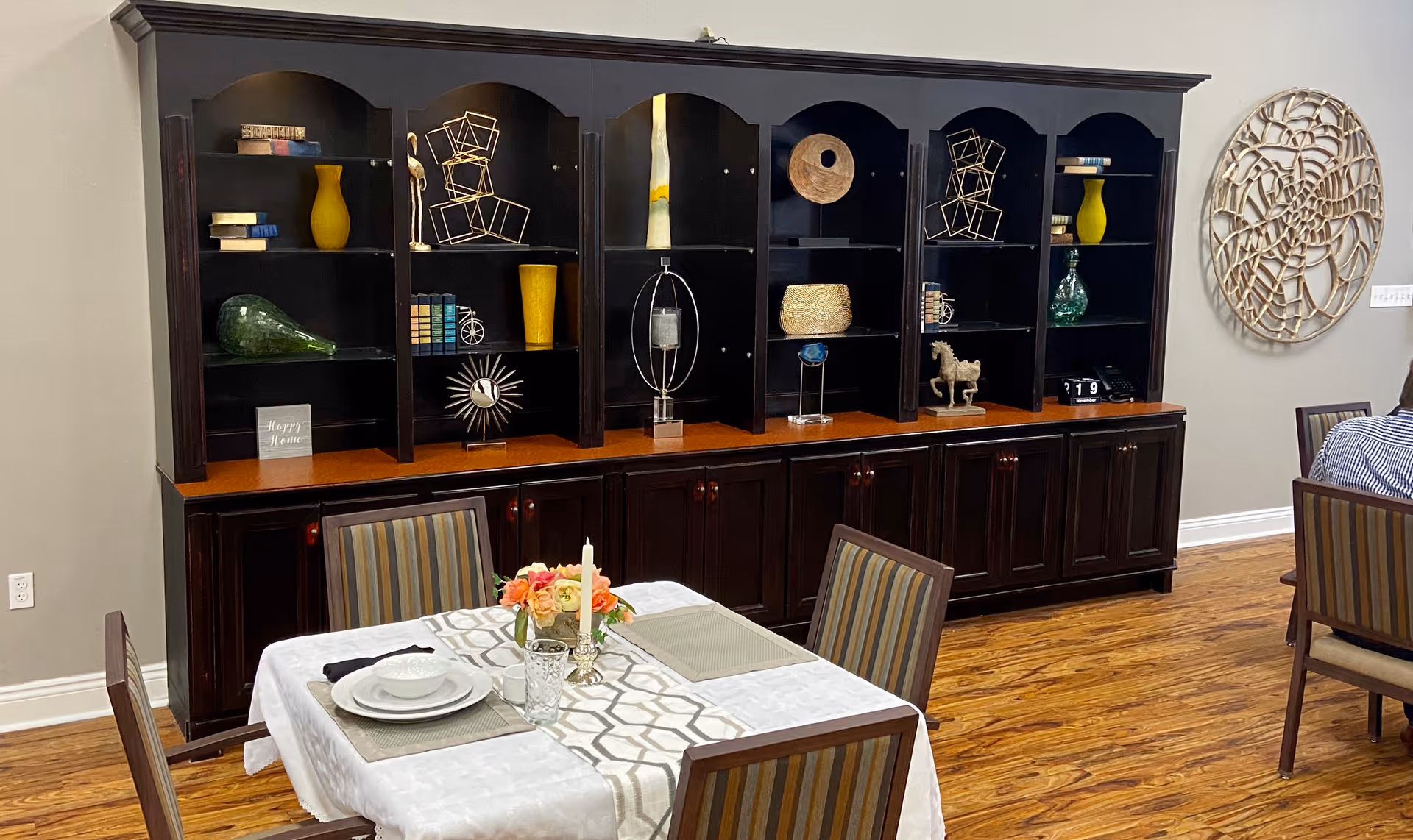 A dining area with a table set for two, featuring white plates, glasses, and a floral centerpiece with candles. Behind the table is a large dark wooden cabinet with shelves displaying decorative items such as vases, books, and sculptures. The room has wooden flooring and neutral-colored walls with a circular wall decoration visible on the right side.